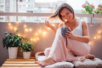 Young women on patio with coffee cup in hand.  There are potted plans and string lights.